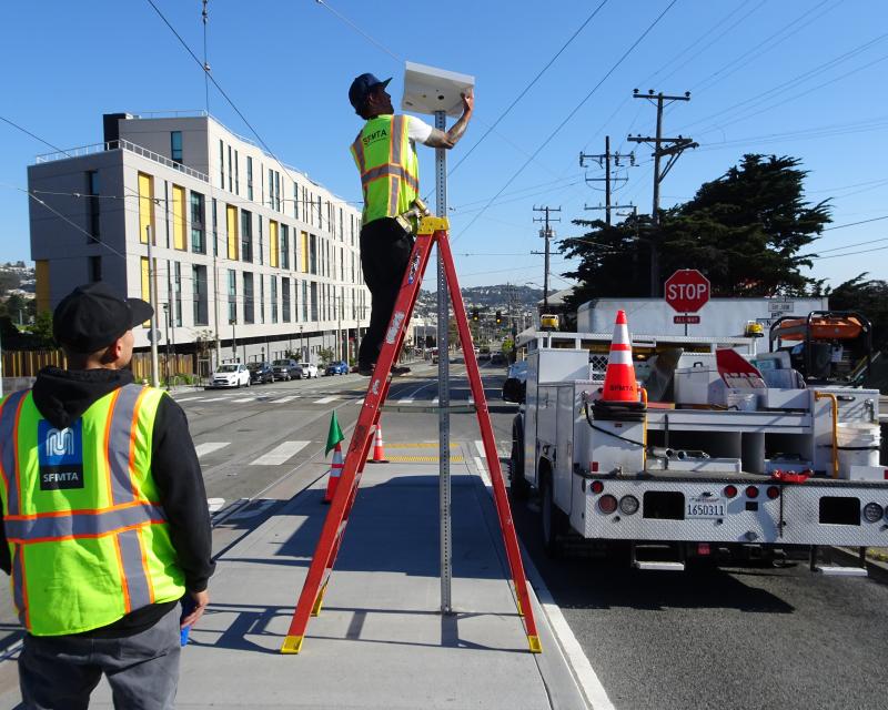 Photo of two SFMTA staff on a boarding platform. One is on a ladder installing a solar panel on top of a pole while another observes.