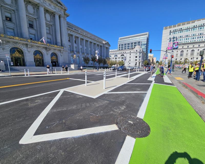 Pedestrians crossing in front of city hall, protected bike lane, painted safety zone, and striped buffer zone all visible in the foreground.