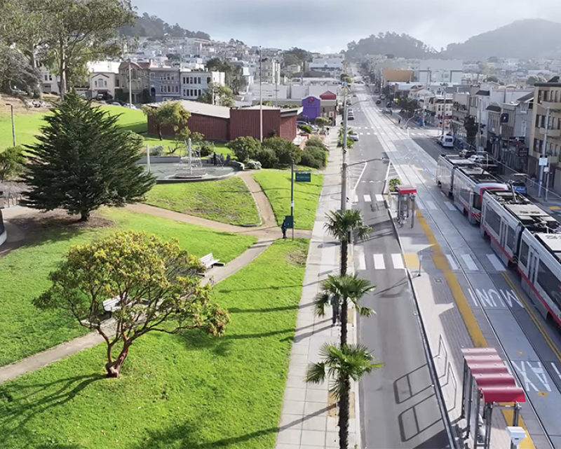 Aerial image of an L Taraval train moving down the street, passing a green space with trees and pathways.