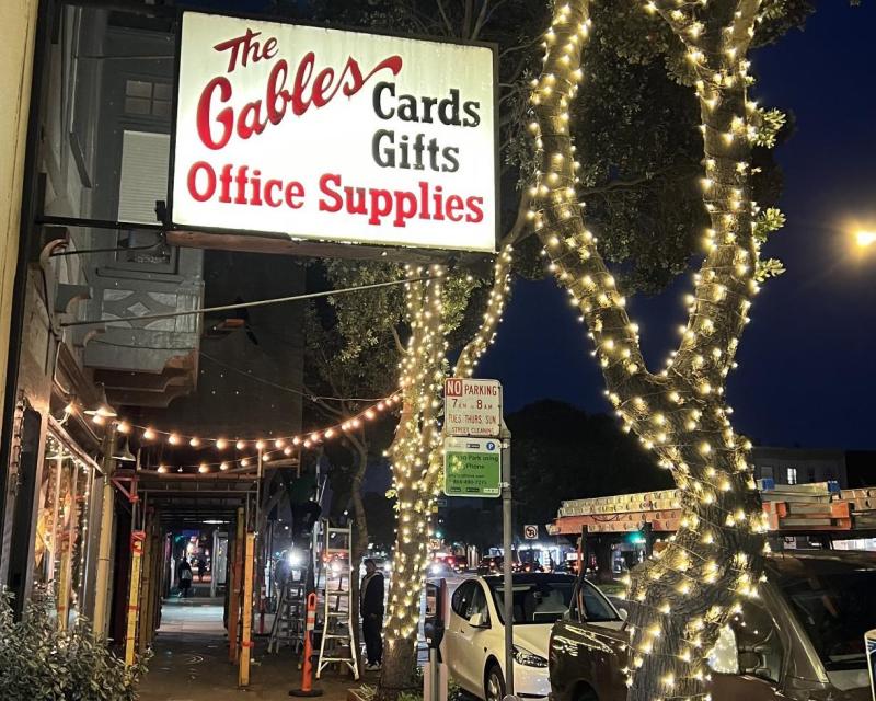 Image shows multiple trees with decorative lighting near the Gables Office Supplies sign on Geary Boulevard.