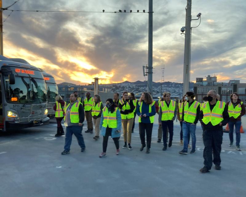 Several people wearing yellow safety vests walk past a Muni bus during a tour of Potrero Yard.