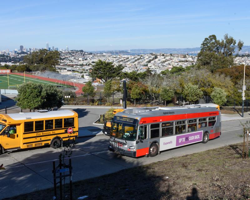 A Muni bus and a school bus use the same street overlooking a school's football field as well as downtown San Francisco.