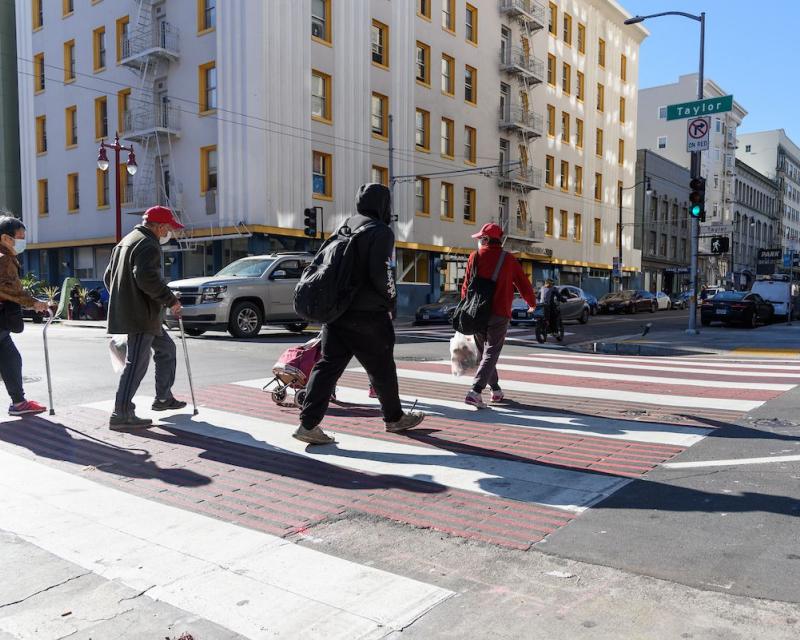 Four people cross a downtown sidewalk painted red and white as cars pass by in the background.