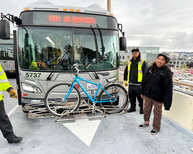 Two people smile in front of a Muni bus that has a bike on the rack.