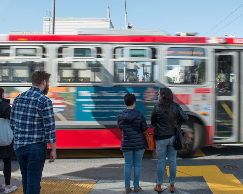 Muni bus passes corner where people are standing.