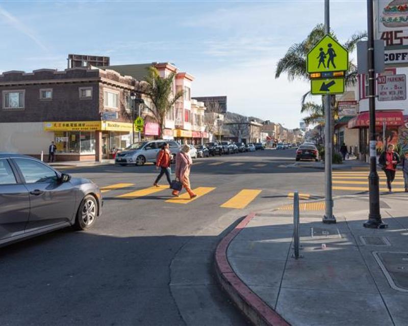 Two people cross a street on a commercial corridor as a car waits.