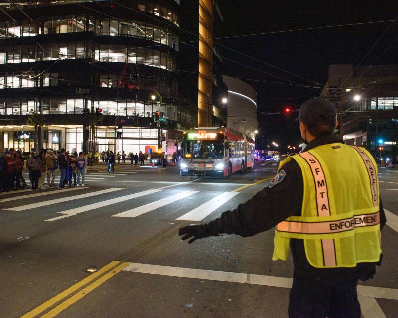 Parking control office wearing a yellow SFMTA vest helps direct traffic as a crowd waits on a corner and a Muni bus approaches.