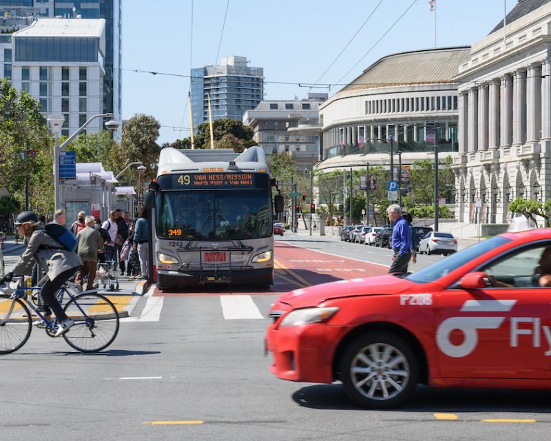 Closeup of an intersection with a person biking, person driving a red cab and bus stopped to pick up passengers.