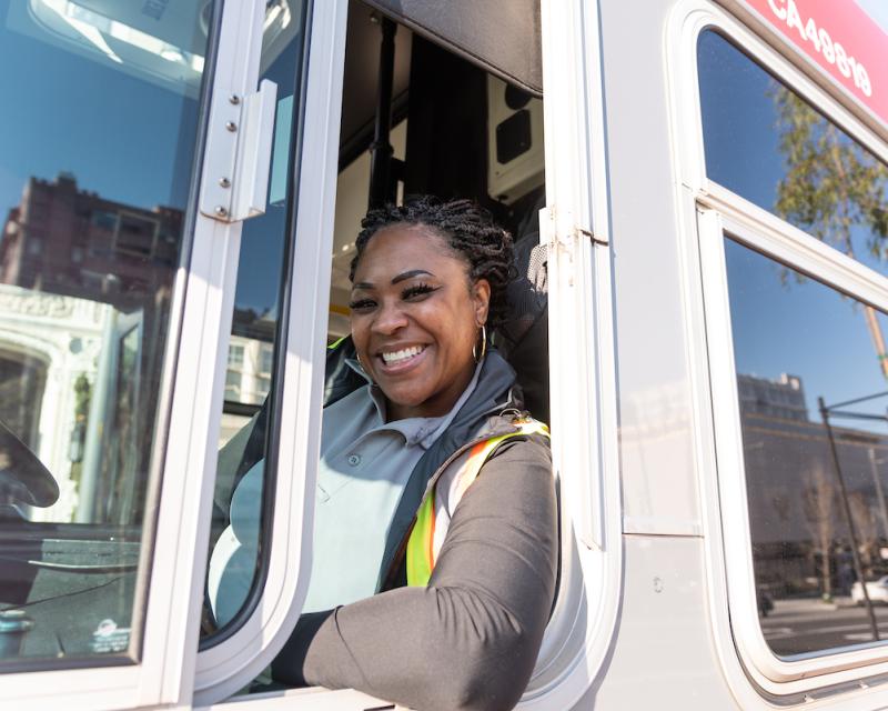 A Muni operator smiles while sitting in the driver seat of a bus.