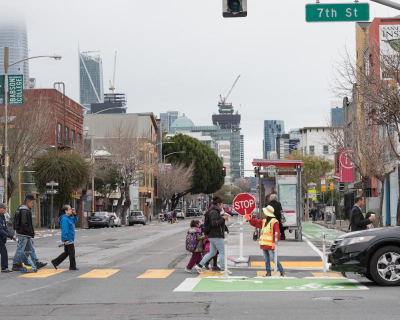 Seniors, children and others cross 7th Street at Folsom with help from a crossing guard who stands in the middle of the crosswalk as a car approaches.