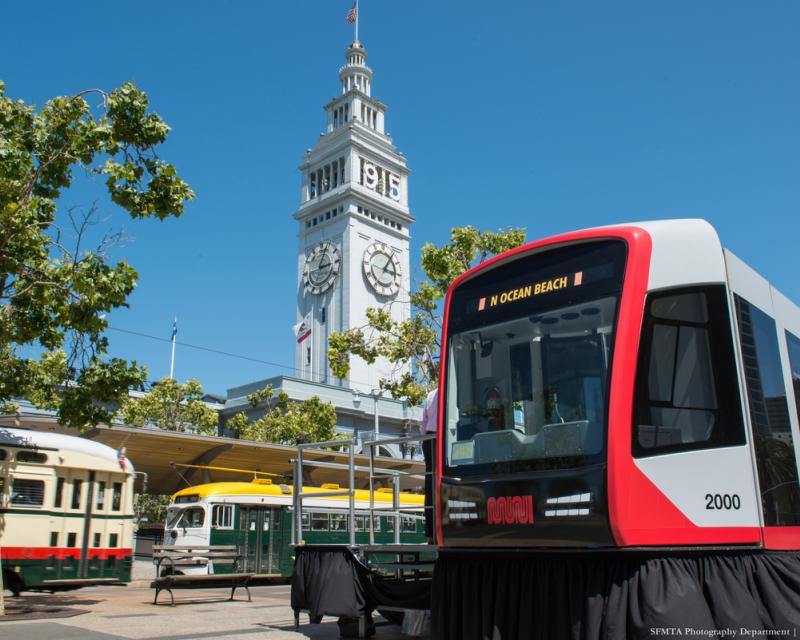 Replica gray and red light rail train model sits on boarding island on The Embarcadero with a blue sky, the Ferry Building and c