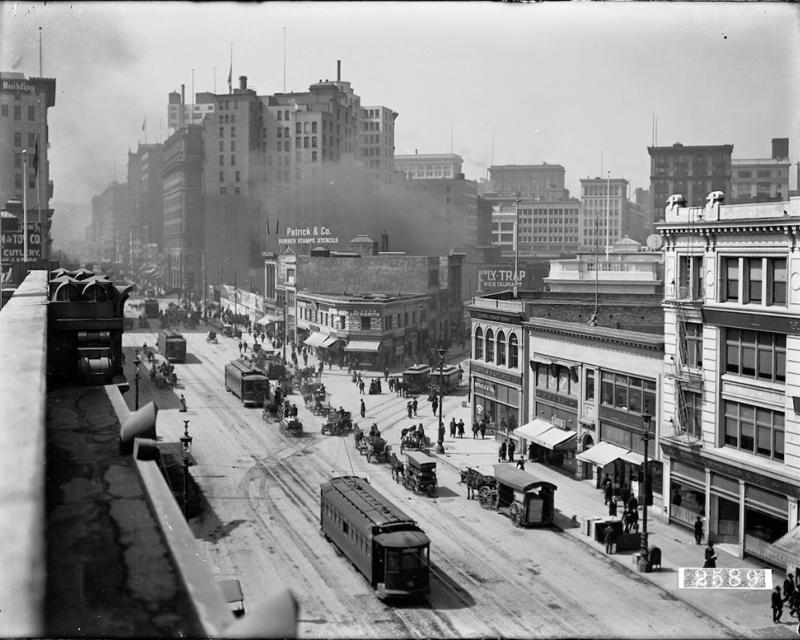 Black and white overhead view of street with horse-drawn and electric streetcars, horse drawn wagons, pedestrians, and buildings
