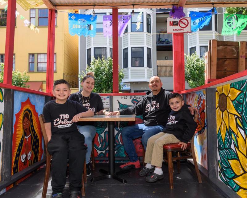 A family shown sitting in a colorful outdoor dining booth. 