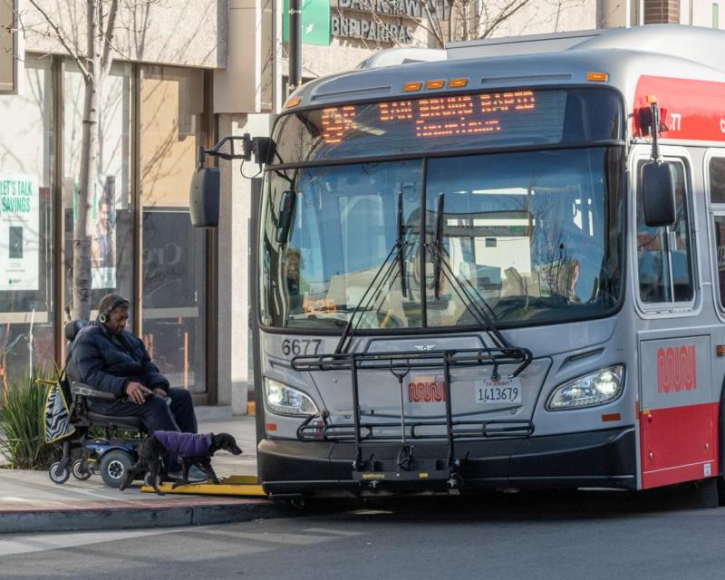 Image of a Muni bus pulled to the curb with its wheelchair ramp deployed as a transit rider using a wheelchair rolls onto bus 