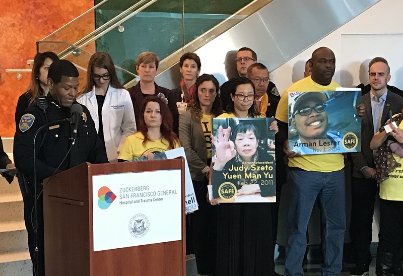 San Francisco Police Chief William Scott speaks at a podium. Behind him are city officials and safe streets advocates wearing yellow shirts and holding large photos of traffic collision victims.