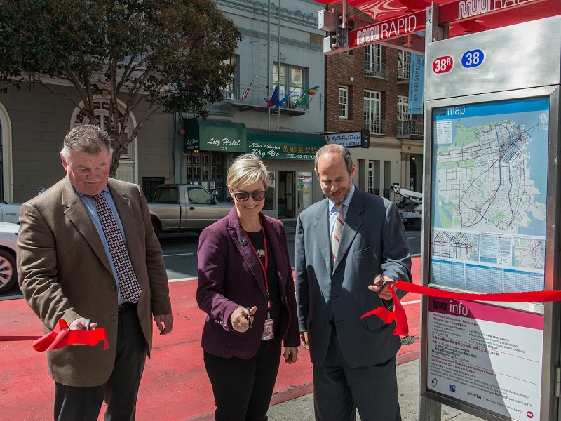 Two men and a woman hold scissors to cut a red ribbon while standing next to a new transit shelter with a red awning and a red traffic lane behind them.