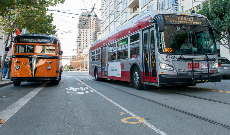 color photo showing two Muni buses next to each other on Steuart street.  On the left is a 1938 orange and black painted White brand bus and on the right is a new 2013 red and grey New Flyer bus.