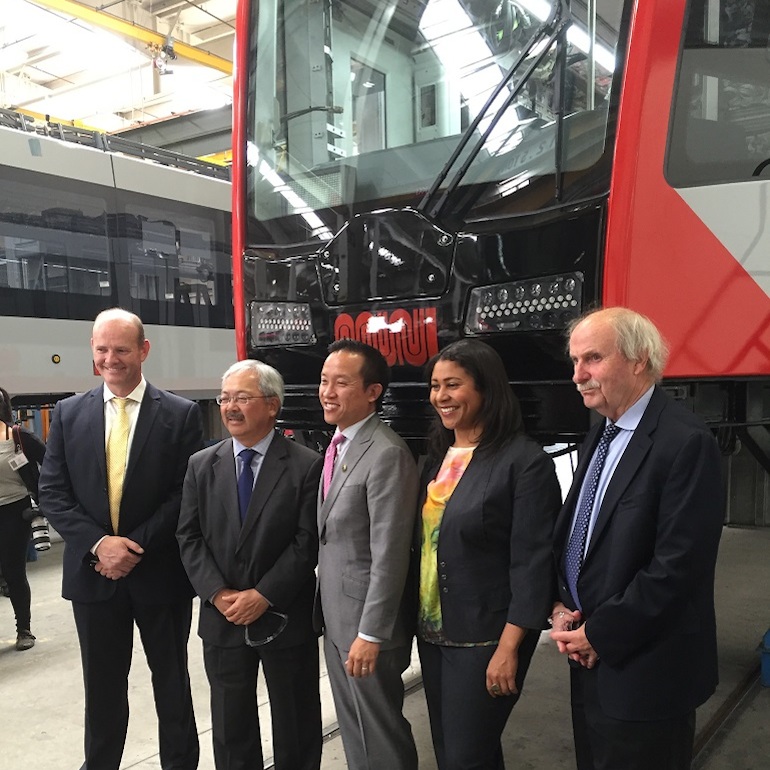 Four men and one woman in suits stand in front of a light rail vehicle on stands, waiting to have the wheel trucks installed.