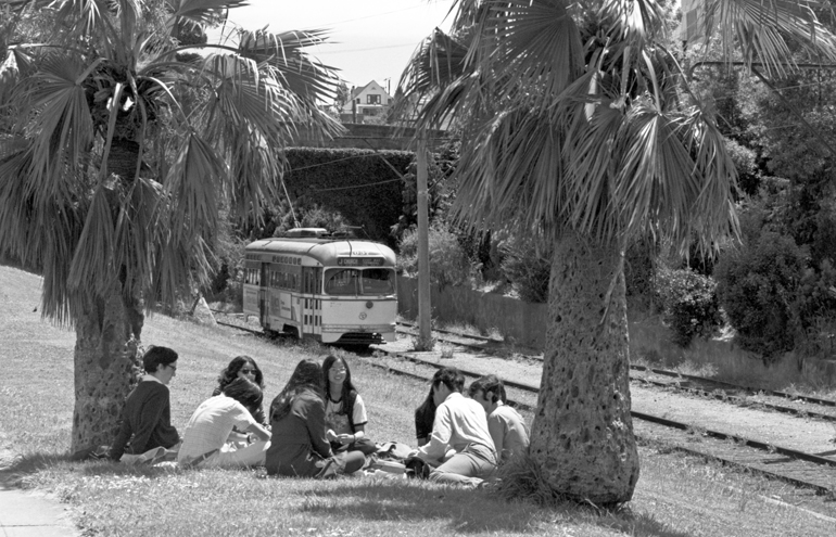 black and white photo from 1972 showing a group of youths sitting in a circle in a park, surrounded by palm trees with a streetcar passing by them in the background