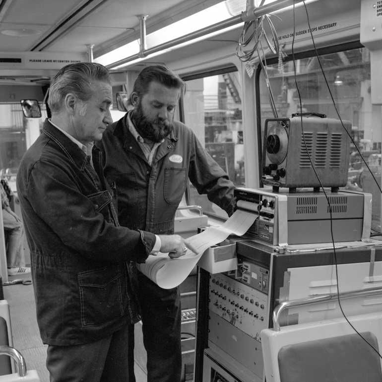 black and white photo showing two men standing in front of large computer inside Boeing Light Rail car looking at graph paper printout coming from machine.