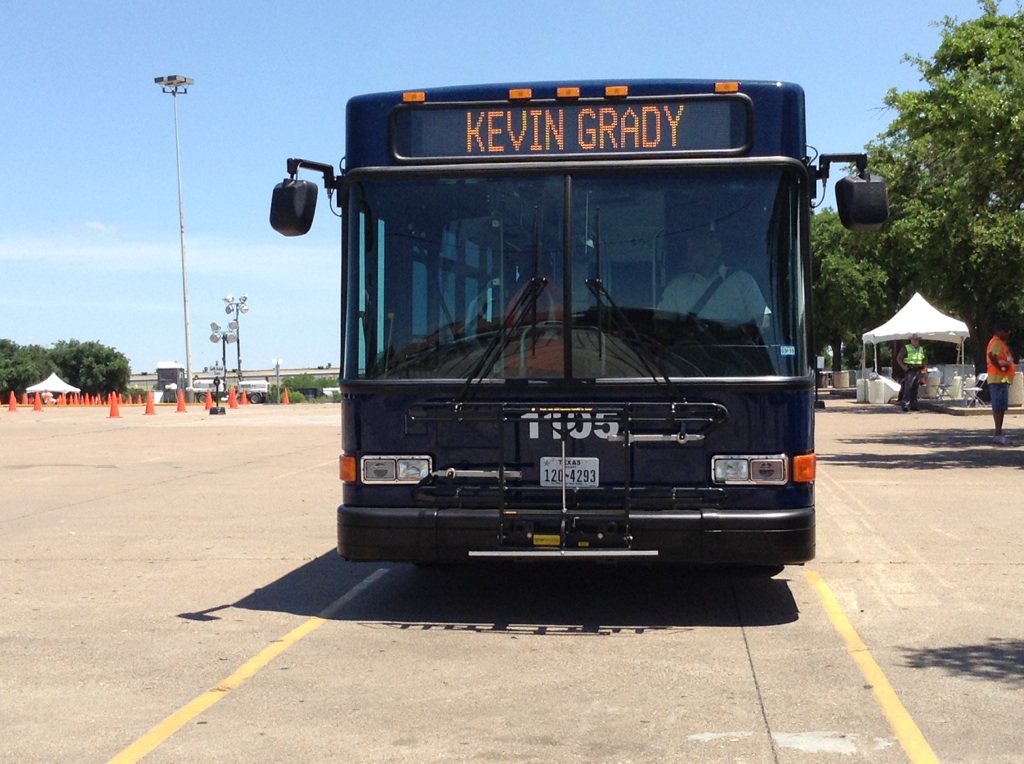 Head sign on a dark blue bus reads "Kevin Grady" as the bus sits in a large, empty parking lot.