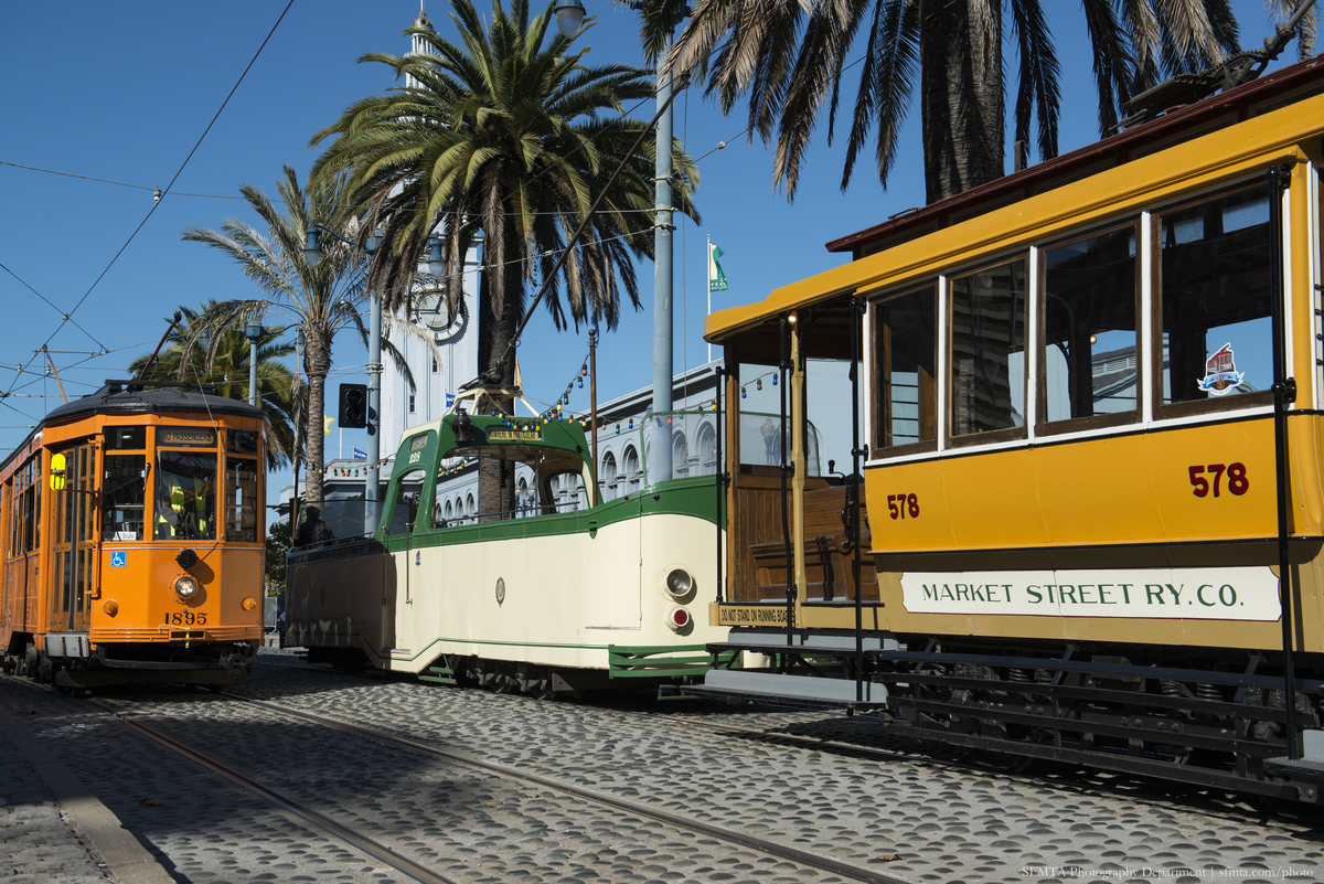Colorful historic streetcars in yellow, green and orange, sit on The Embarcadero with a blue sky overhead.