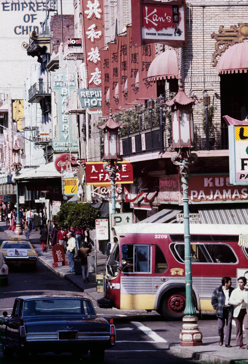 Colorful street scene of Chinatown in 1972 with a maroon and yellow Muni bus crossing the street.